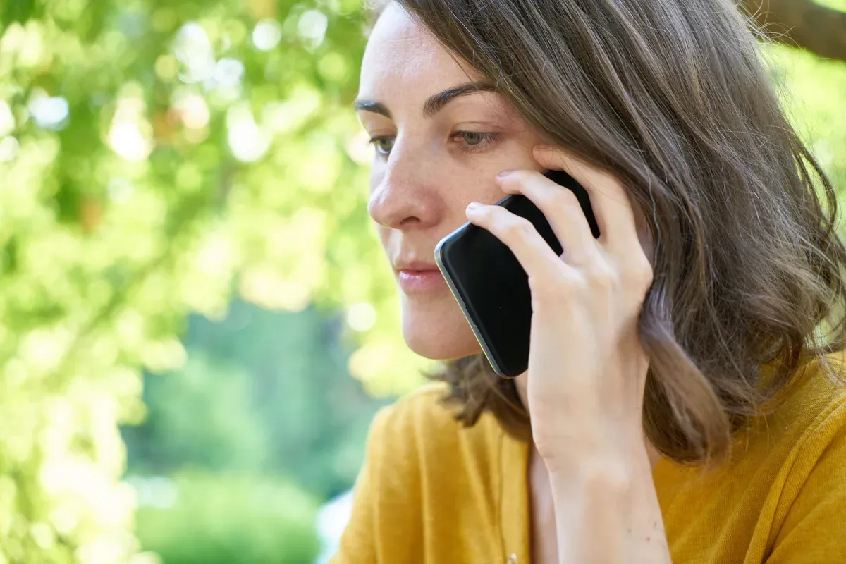 mulher de blusa amarela falando ao telefone com expressão séria, ao ar livre