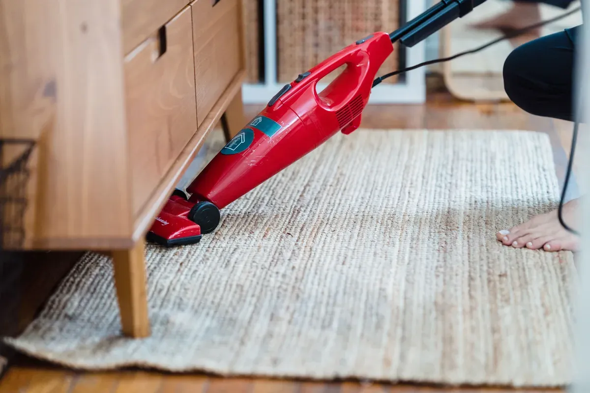 aspirador de pó vermelho passando num tapete de fibra natural, com a ponta encostada na lateral de um móvel de madeira